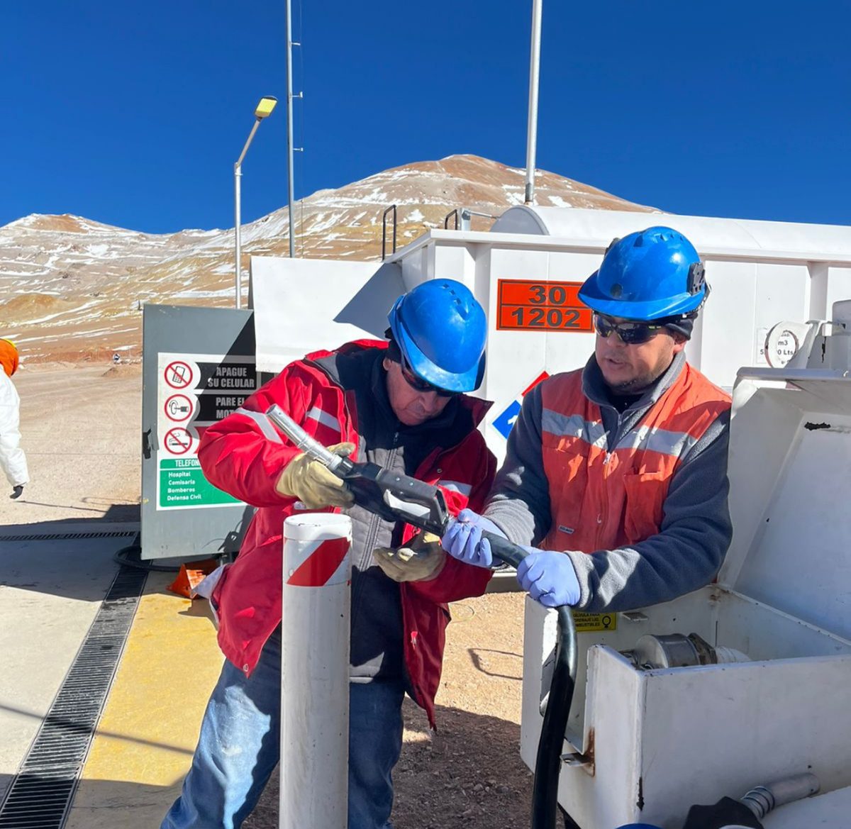 Operarios de Surmat con ropa térmica y arnés trabajando sobre un equipo de despacho de combustible en una planta de alta montaña, con la cordillera nevada de fondo